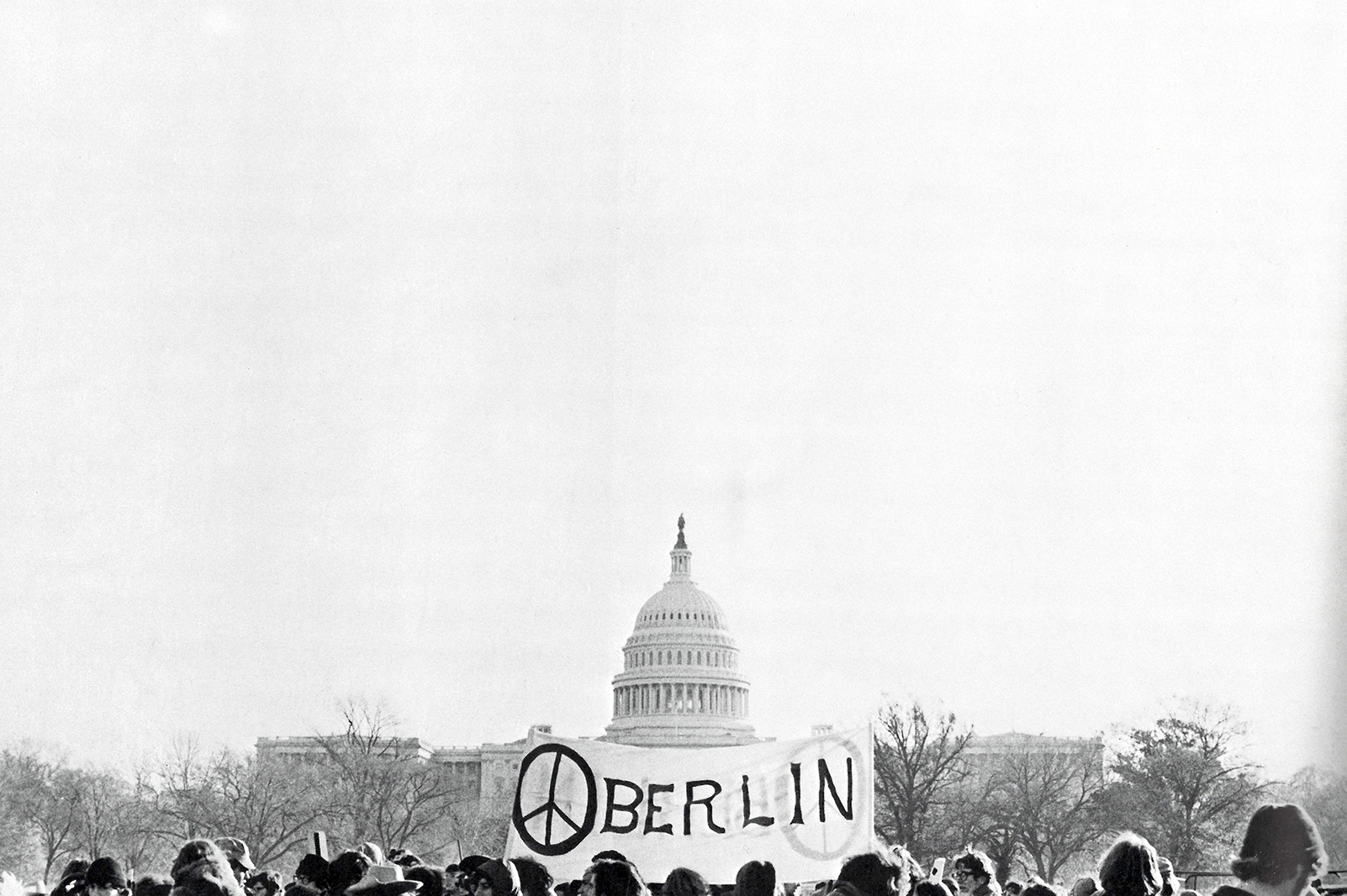 A crowd gathers by the U.S. Capitol, holding a banner that says 'Oberlin' with the letter O containing the peace symbol. Black and white archival photo.