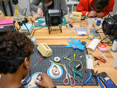 A group of students work at a table filled with shapes, wires, and cutting tools.